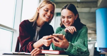 Two friends smiling while looking at a cell phone.