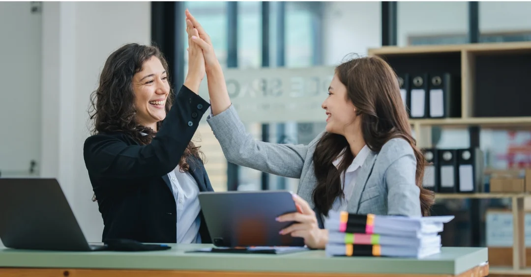 Two co-workers at a desk giving each other a high-five.