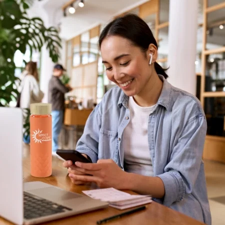 Smiling person wearing ear buds, sitting at a desk and looking at a smartphone.