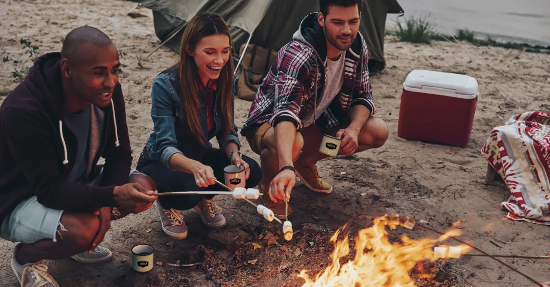 Three campers roasting marshmallows over a campfire while resting near the lake.