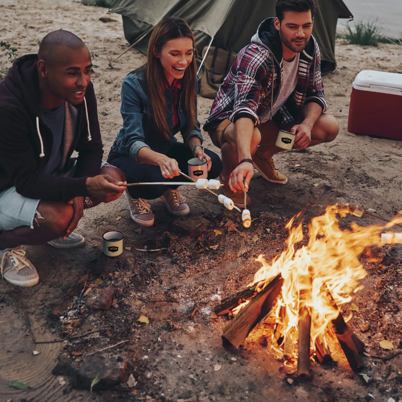 Three campers roasting marshmallows over a campfire while resting near the lake.