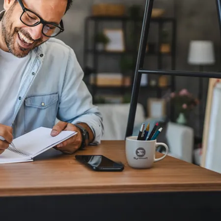 Smiling person writing in a notebook. A mug filled with pens sits on his desk.