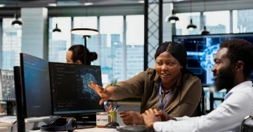Boss explaining onboarding to new employee at desk in front of computer monitors