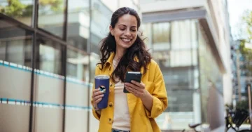 Person in a yellow jacket walks down the street holding a travel coffee mug and phone.