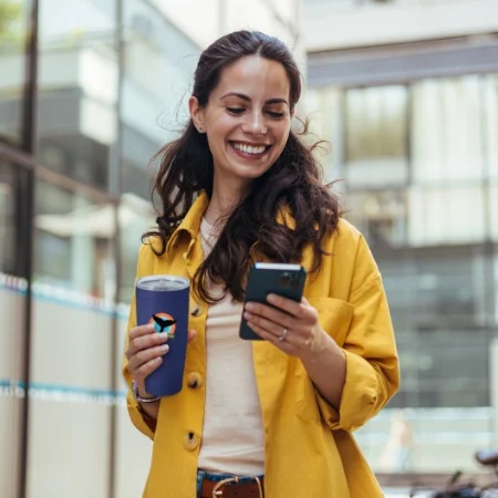 Person in a yellow jacket walks down the street holding a travel coffee mug and phone.