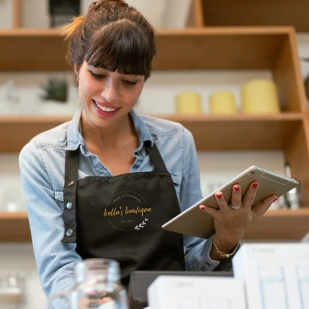 Smiling boutique owner is wearing an apron and holding a tablet. 