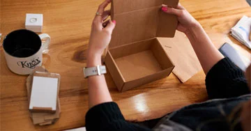 person assembling a small, open, brown cardboard box on a wooden table