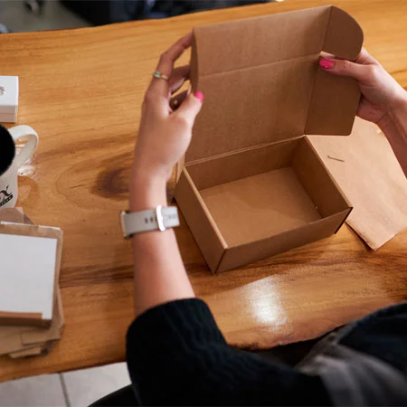 person assembling a small, open, brown cardboard box on a wooden table