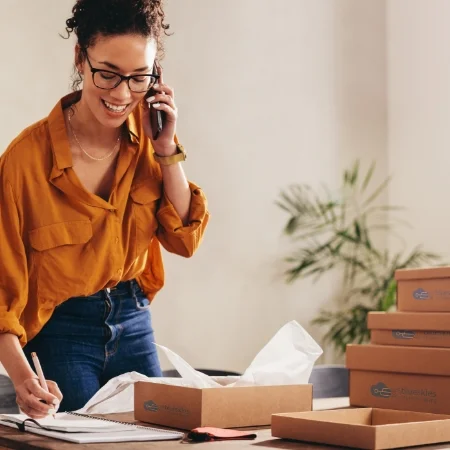Business owner on the phone. Stacked boxes are on a table next to them. 
