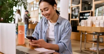 Smiling person wearing ear buds, sitting at a desk and looking at a smartphone. 