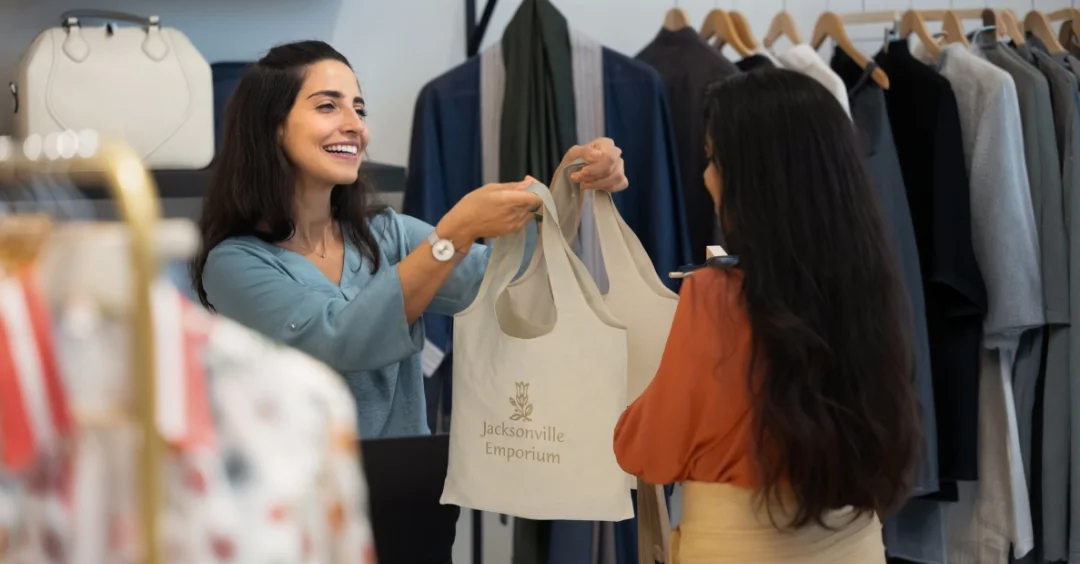 Smiling store clerk handing customer their purchase in a reusable branded shopping tote.