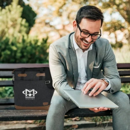  Person with a laptop sitting on a bench in a park with their laptop case next to them.