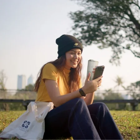 Woman sitting outside with a beanie on and reusable bag by her side while drinking out of a reusable mug