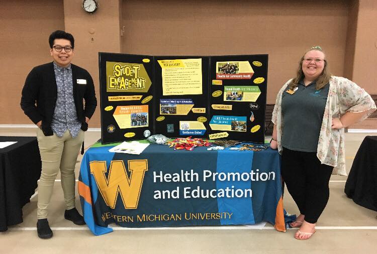 Customer photo from Jeremy of our Serged Closed-Back Table Throw - 8' - Full Color, a couple of people standing next to a table with a table with a sign