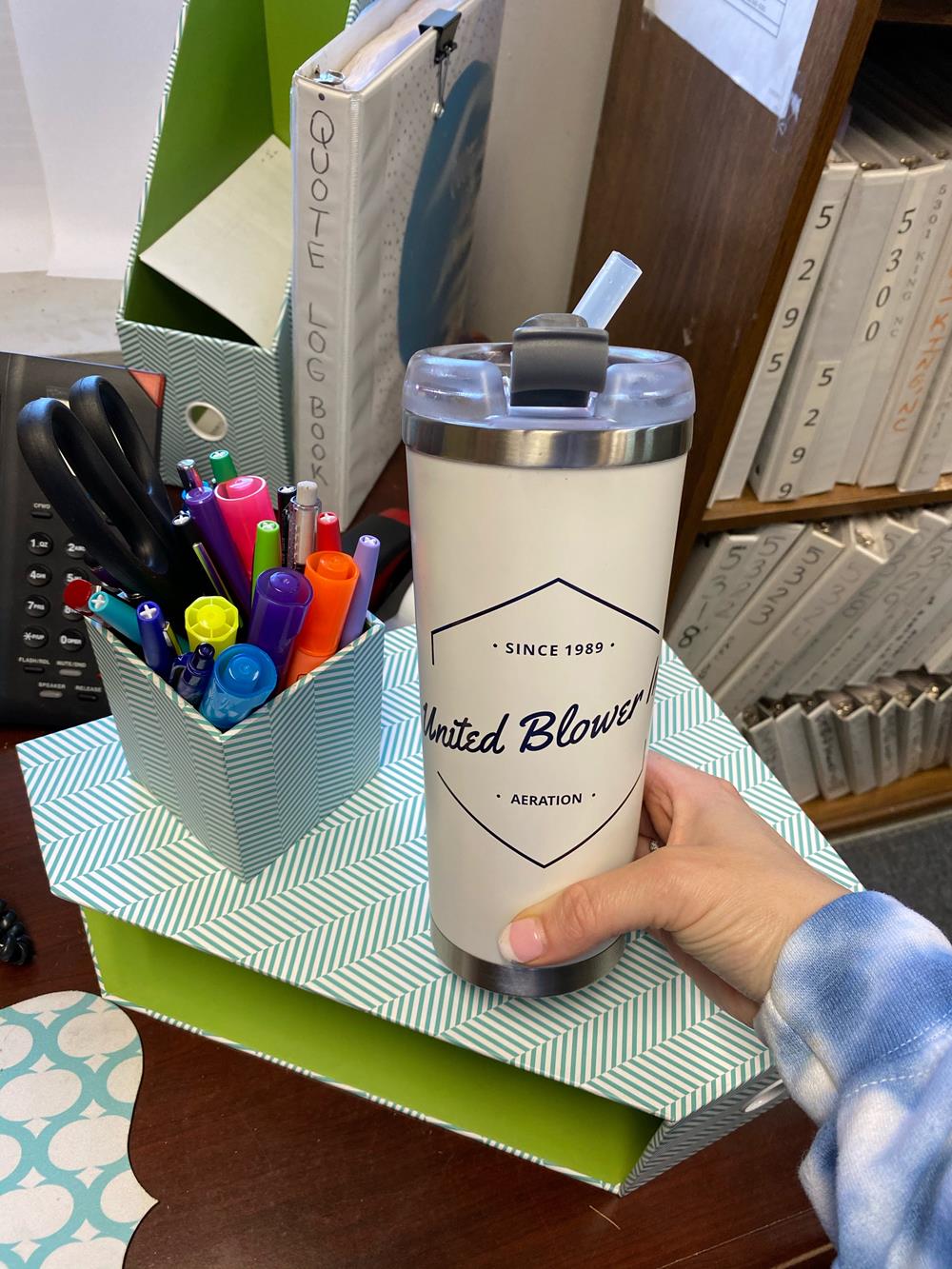 a hand holding a white and silver tumbler with a straw on a green box with pens and a green box with a blue and white striped box with a blue and white striped box with a black