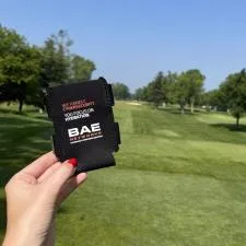 Customer photo from Sarah of our Koozie® Duo Can Cooler, a hand holding a black object in front of a golf course